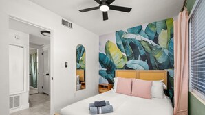 A bedroom with a bed, a ceiling fan, a bathroom with a shower curtain, and a wall with a tropical leaf mural. - Beautiful East Orlando Home Near Everything (Orlando)