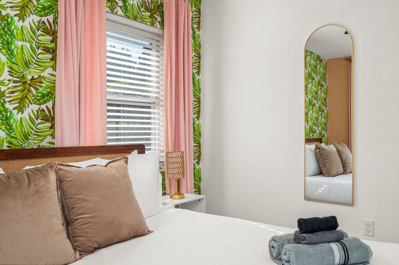 A bedroom with a bed, a bedside lamp, a mirror, and a window with blinds.