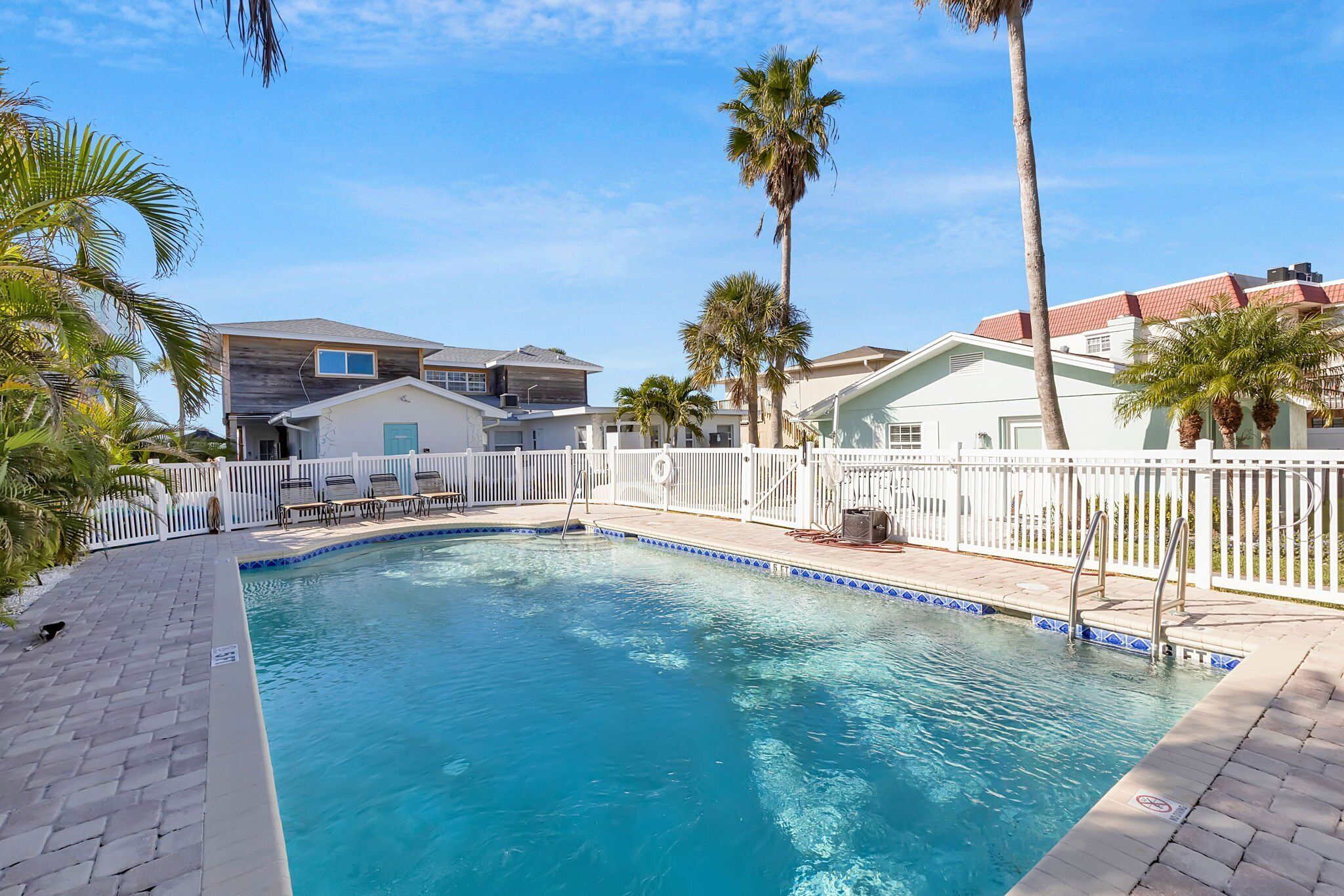 A swimming pool with a white fence, palm trees, and houses in the background.