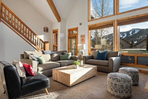 A spacious living room with a wooden staircase, large windows with mountain views, and a cozy seating area with a coffee table and ottomans. - Lakota Antlers 108 | Private Hot Tub | Slope Views (Winter Park)