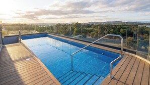 A rooftop pool with clear blue water, surrounded by a wooden deck and glass railings, offering a view of distant hills and trees. - Stylish 1BDR Sunlit Apartment w Rooftop Pool and Gym (Canberra)