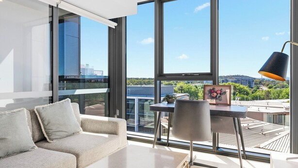 A modern living room with a large window, a sofa, a table, and a lamp. - Stylish 1BDR Sunlit Apartment w Rooftop Pool and Gym (Canberra)