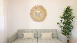 Een lichtgrijze bank met twee kussens, een ronde spiegel met franjes, en een hoge groene plant ernaast.