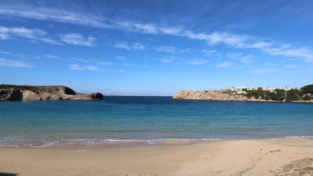 Una playa con agua azul cristalina, orilla de arena, afloramientos rocosos y edificios a la derecha.