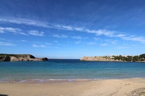 A beach with clear blue water, sandy shore, rocky outcrops, and buildings on the right.