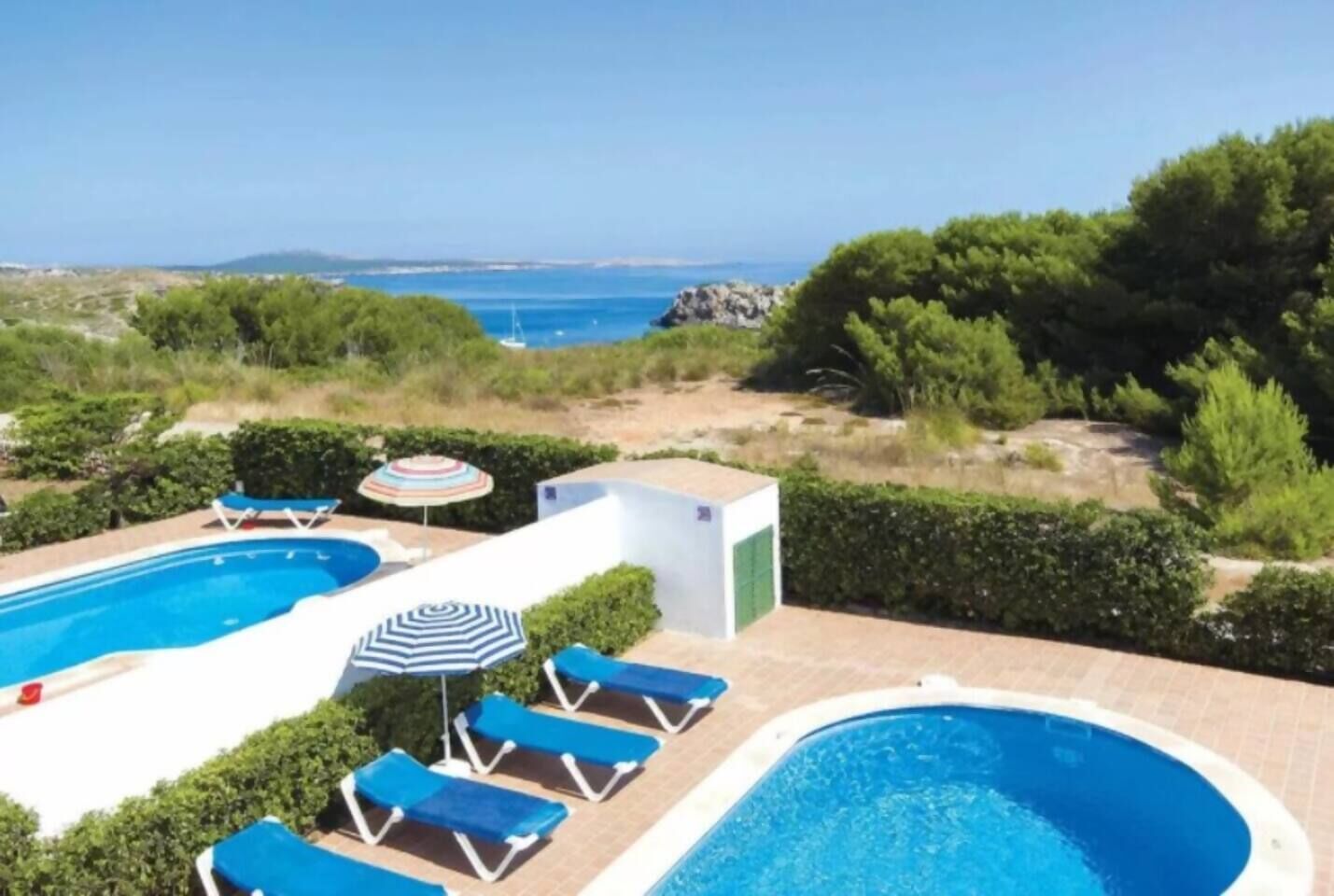 A swimming pool with lounge chairs and umbrellas, surrounded by greenery and a view of the sea.