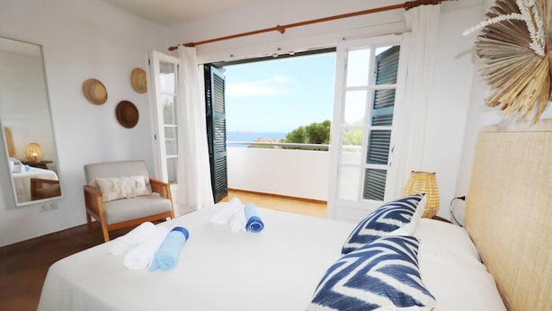 A hotel room with a bed, a chair, a small table, and a view of the sea through open shutters. - Villa Las Marinas 3, pool and 5 minutes to the sea (S'Arenal D'En Castell (Menorca) (Urbanitzacio))