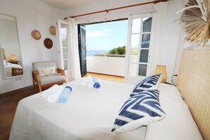 Et hotelværelse med seng, stol, lille bord og havudsigt gennem åbne skodder.