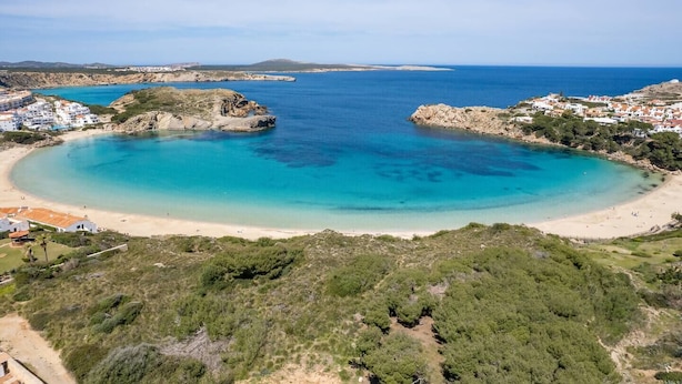 Kystområde med strand, klart blåt vand og omkringliggende bygninger.