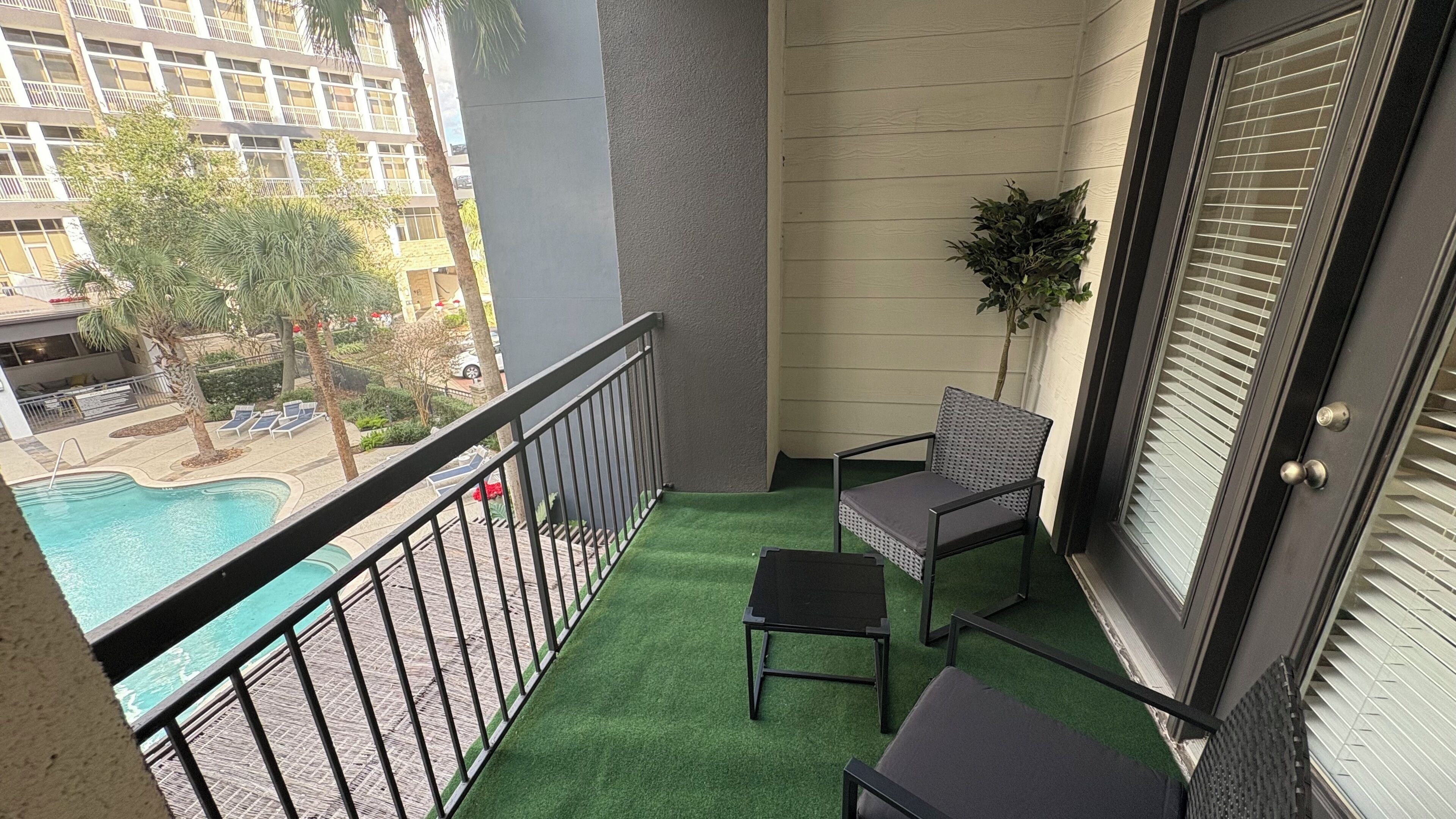 A balcony with artificial grass, a chair, a small table, a potted plant, and a view of a swimming pool and buildings.
