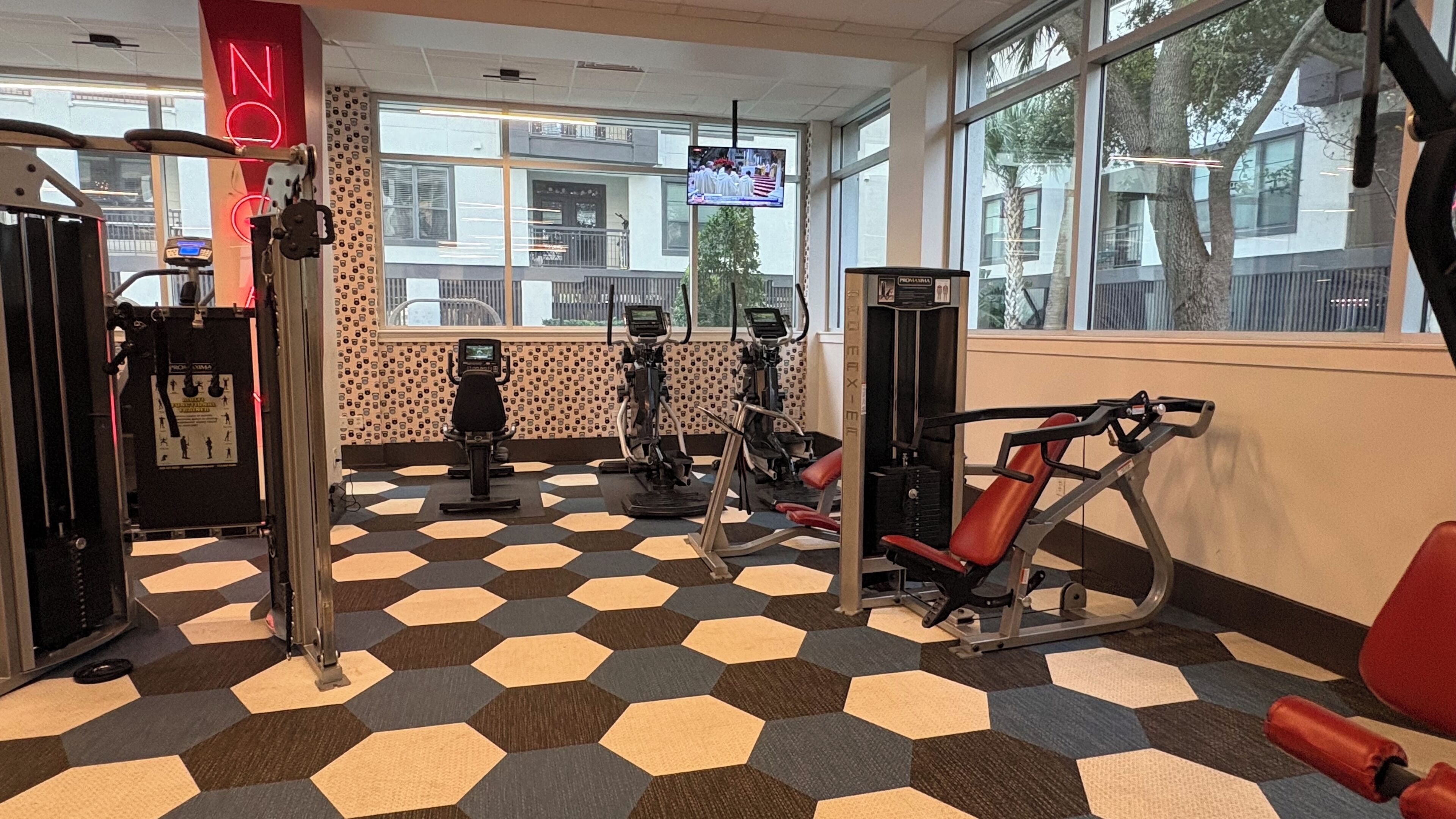 A gym with various exercise machines, a patterned floor, and large windows with a view of the outside.