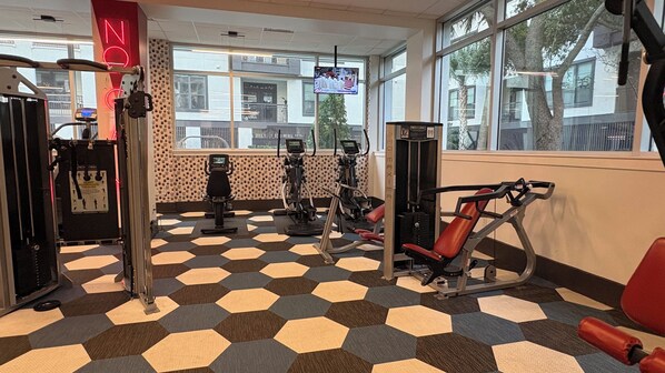 A gym with various exercise machines, a patterned floor, and large windows with a view of the outside.