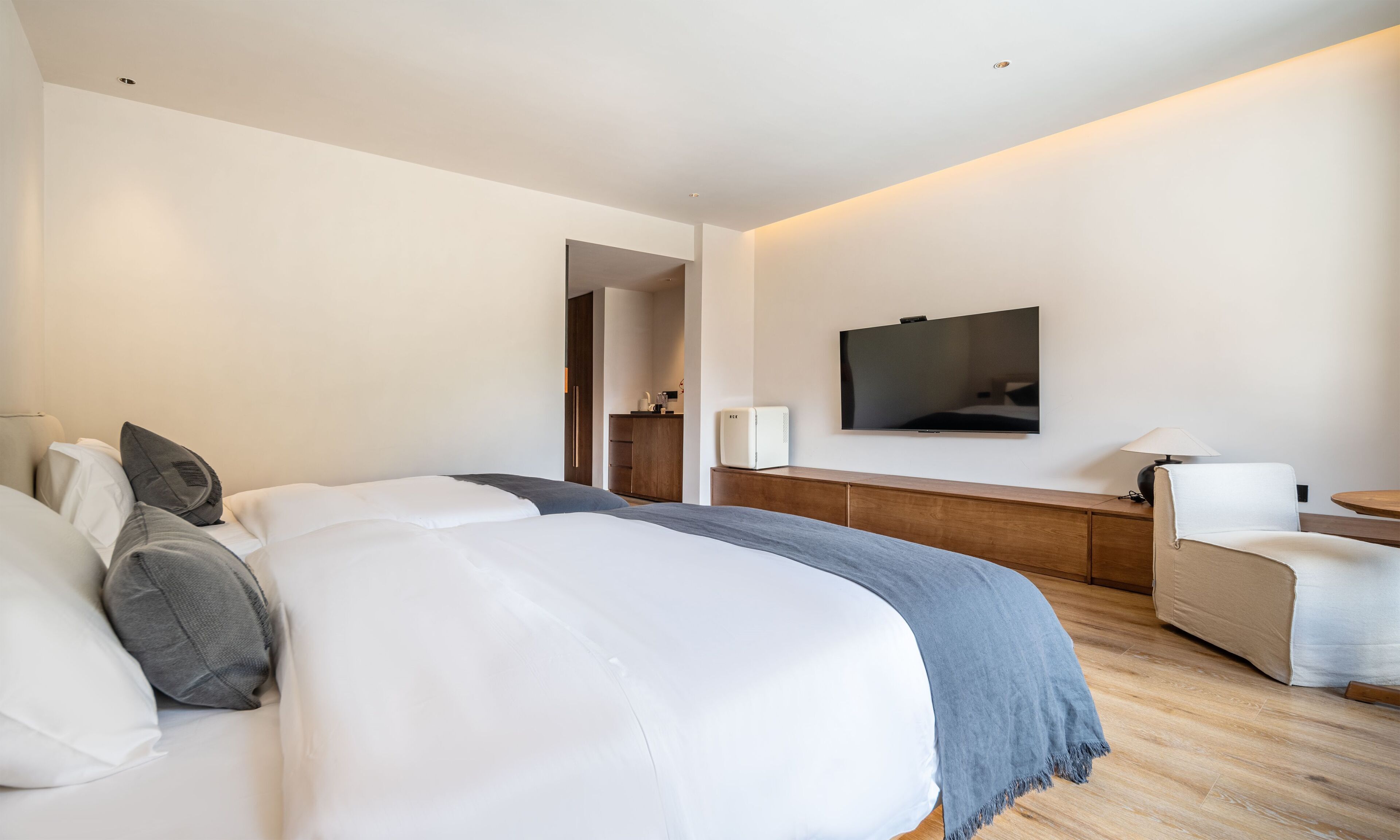 Una habitación de hotel moderna con una cama grande, un televisor montado en la pared, un escritorio con una lámpara y una pequeña kitchenette al fondo.