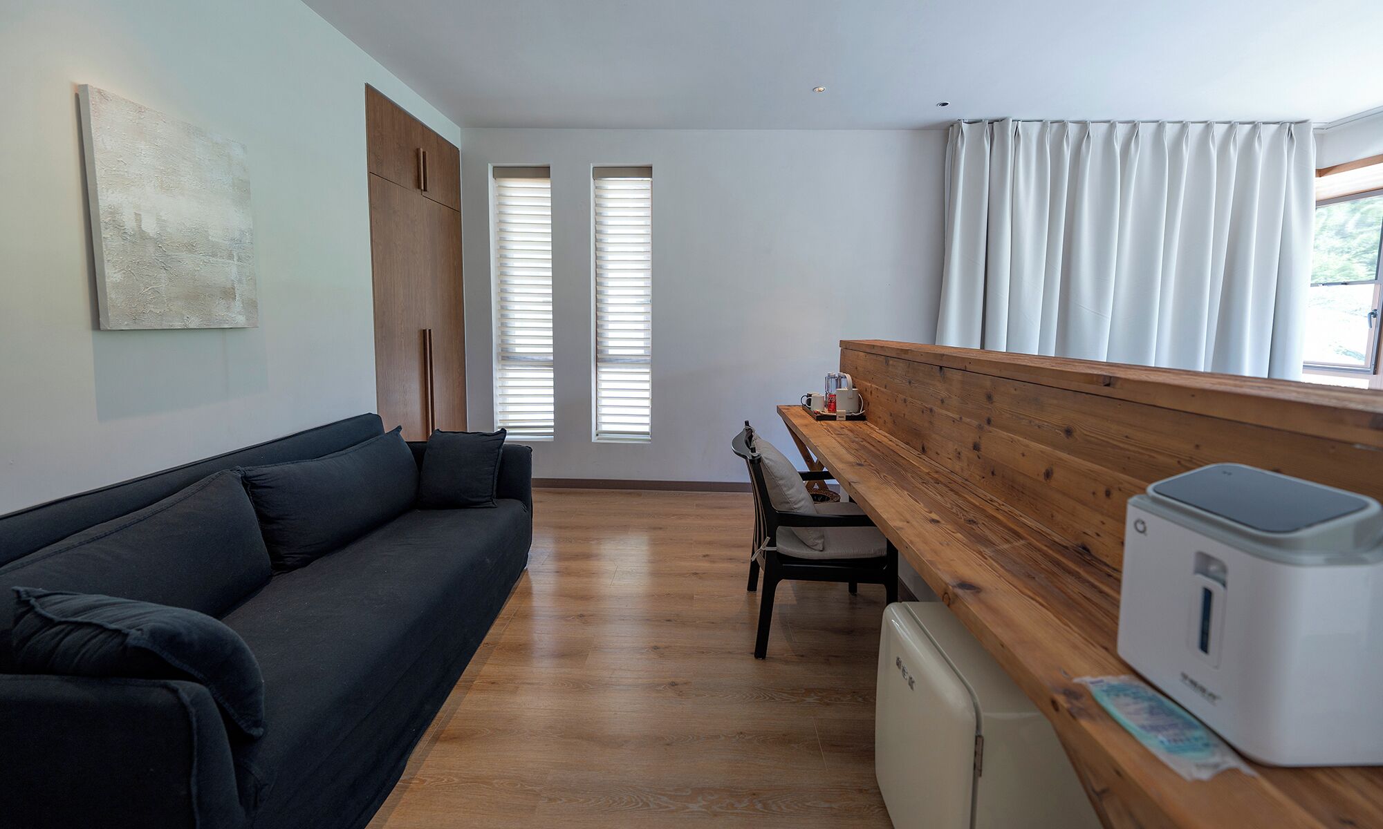 A modern living room with a dark sofa, wooden bench, and a small table with a kettle. There are two windows with blinds, a white wall, and a ceiling light.