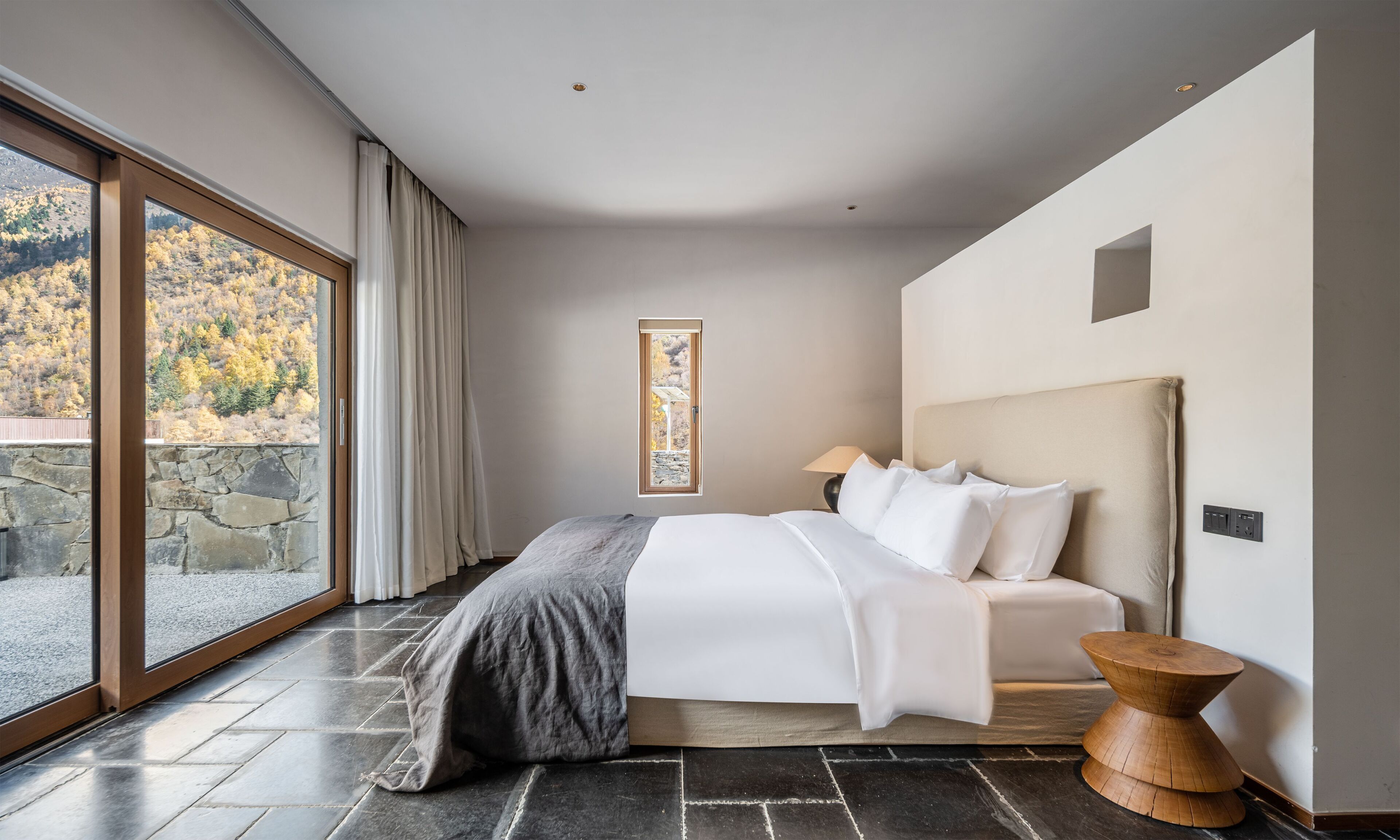 A modern bedroom with a large window, a bed with white linens, a stone wall, and a wooden side table.