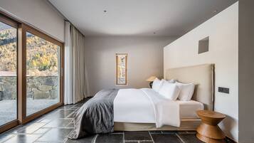 A modern bedroom with a large window, a bed with white linens, a stone wall, and a wooden side table.