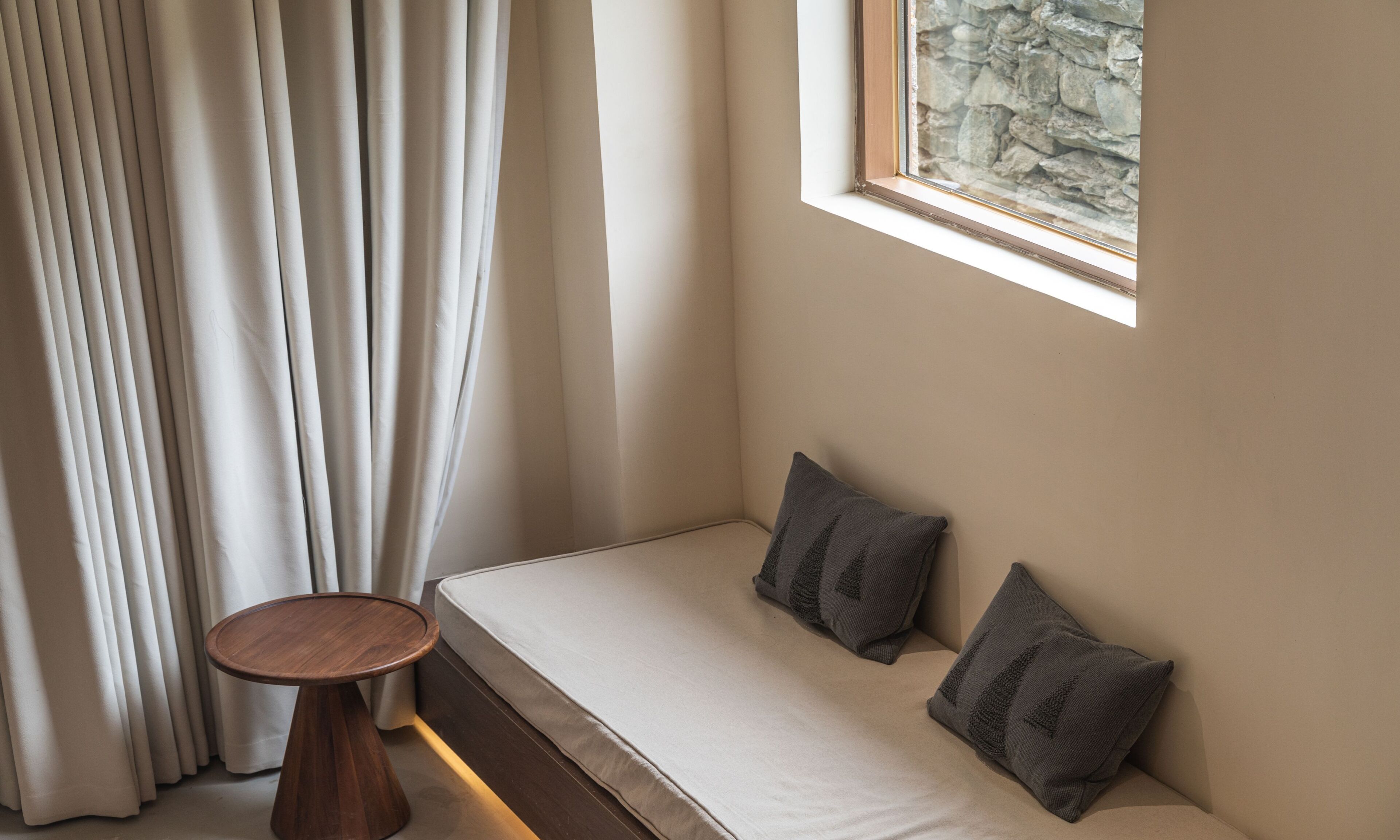 Habitación de esquina con cama, una mesita de madera y ventana con vista a un muro de piedra.