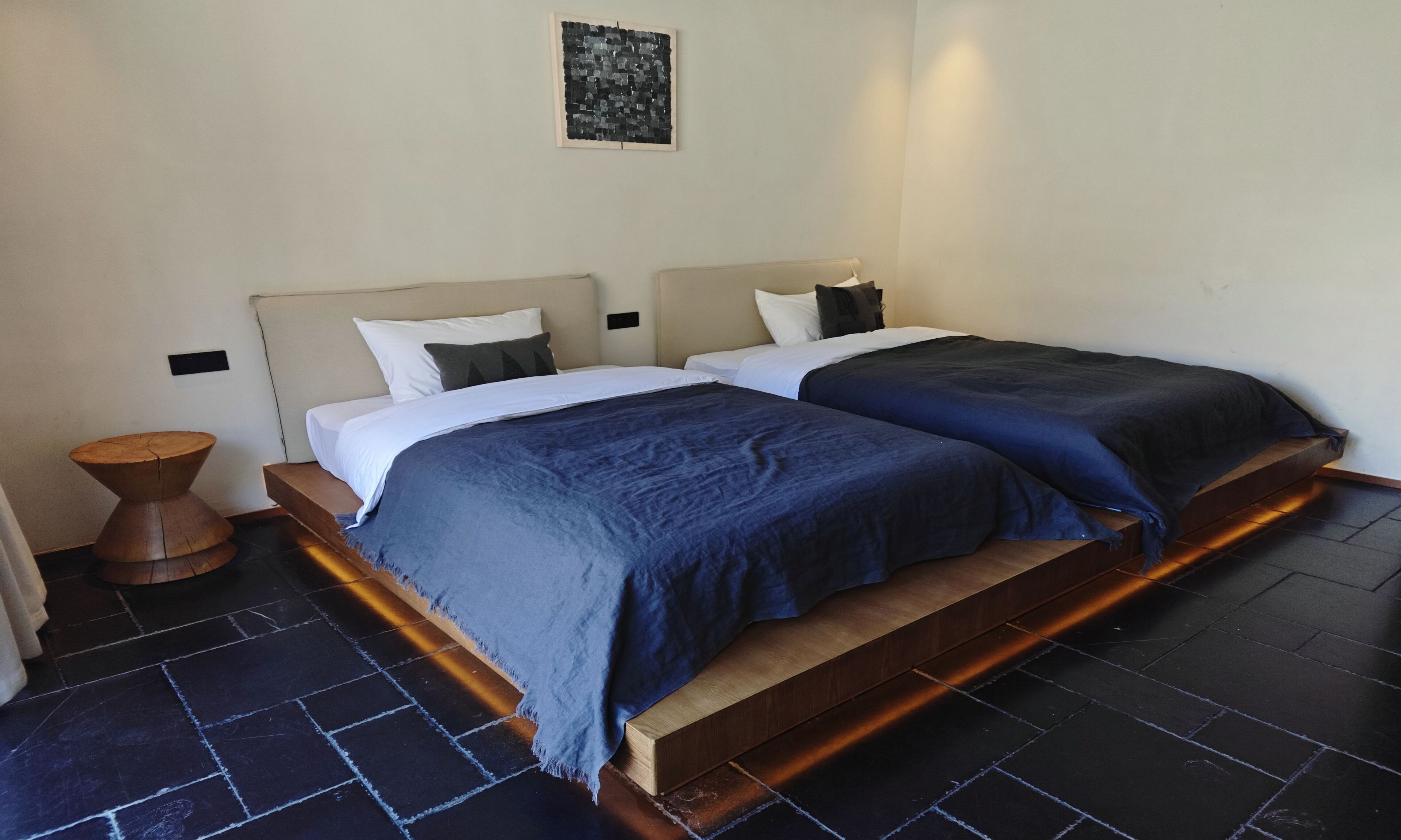A double bed with a wooden platform, a small wooden stool, and a wall-mounted light fixture.