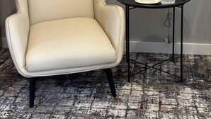 Un sillón color crema con patas negras, una mesita redonda con un objeto blanco encima y un enchufe en la pared.