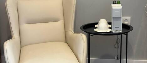 Un sillón color crema con patas negras, una mesita redonda con un objeto blanco encima y un enchufe en la pared.