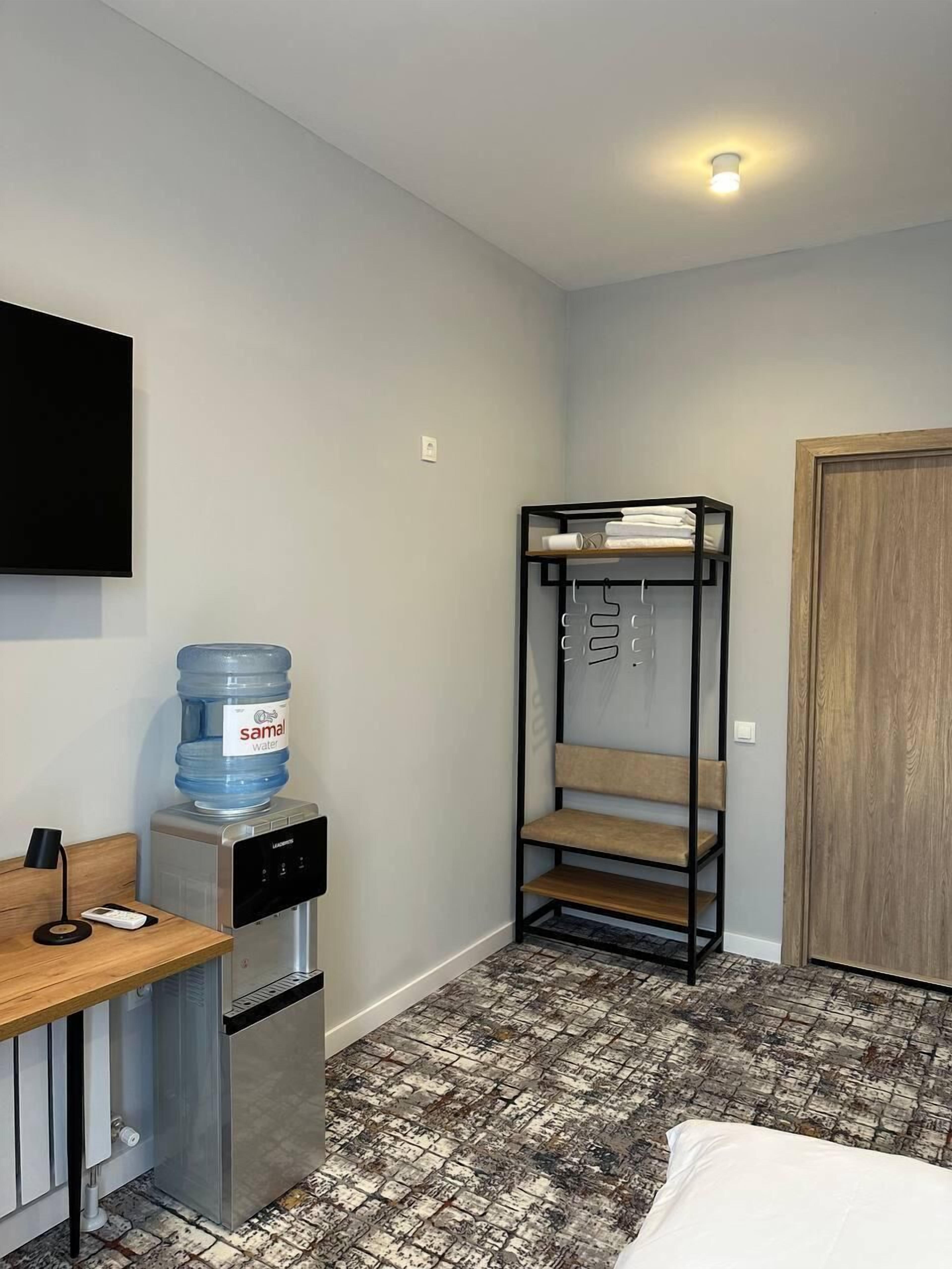 A compact hotel room with a kitchenette, a water dispenser, a bench with hanging items, and a wooden door.