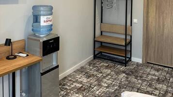 Habitación de hotel compacta con kitchenette, dispensador de agua, un banco con objetos colgando y una puerta de madera.