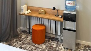 Habitación de hotel con televisión de pantalla plana montada en la pared, un escritorio de madera con dispensador de agua, una cafetera y un taburete pequeño.