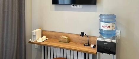A hotel room with a flat-screen TV mounted on the wall, a wooden desk with a water dispenser, a coffee machine, and a small stool.