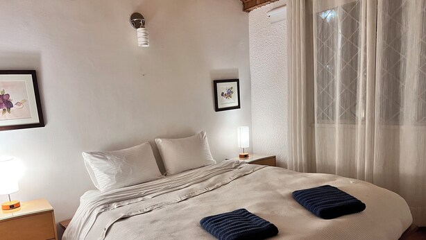 Ein ordentlich bezogenes Bett mit einer weiß-blauen Streifendecke, ein Nachttisch mit einer Lampe und ein gerahmtes Bild an der Wand.