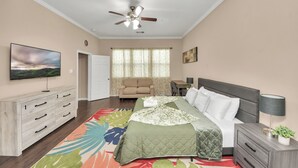 Un dormitorio con cama, mesita de noche, televisor, ventilador de techo y una ventana con cortinas.