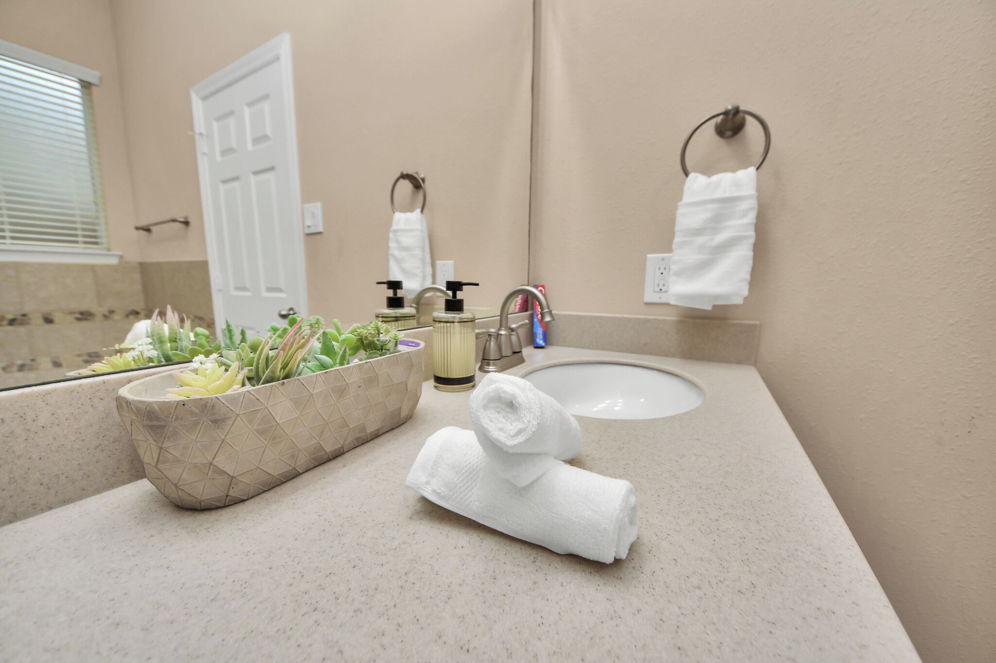 Une salle de bain avec un comptoir beige, un lavabo, un miroir, une serviette, un distributeur de savon et une plante en pot.