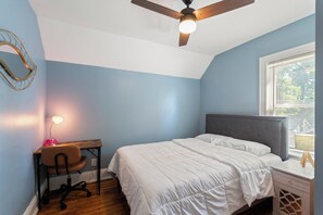 Quarto com cama, escrivaninha, cadeira, ventoinha no teto e janela.