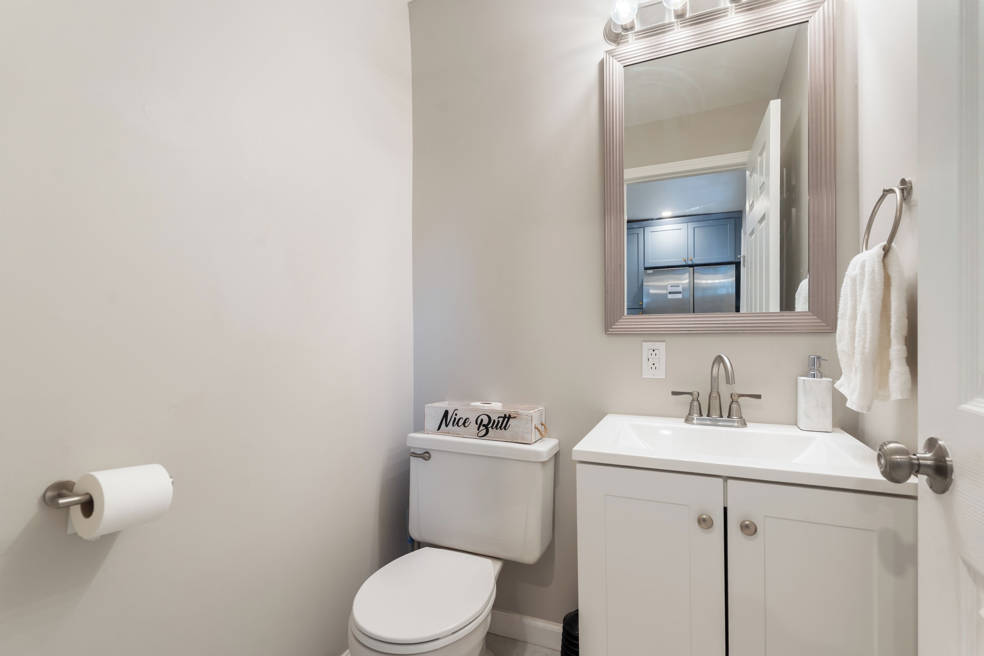 A bathroom with a toilet, sink, mirror, and a box labeled 'Nice Butt' on the toilet tank.