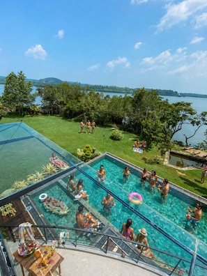 Una piscina con agua cristalina, rodeada de césped y árboles, con vista a un lago y colinas al fondo.