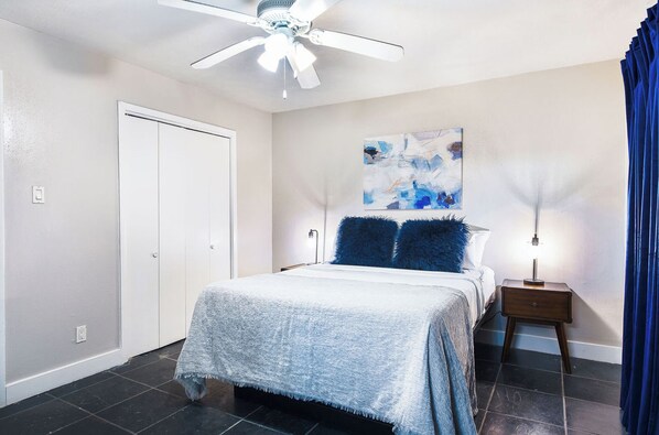 Une chambre à coucher avec un lit, un ventilateur de plafond, un tableau, une table de chevet et un rideau.