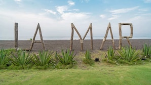 Letras de madera que deletrean "LAMAR" en una playa, con pasto y plantas de agave al frente.