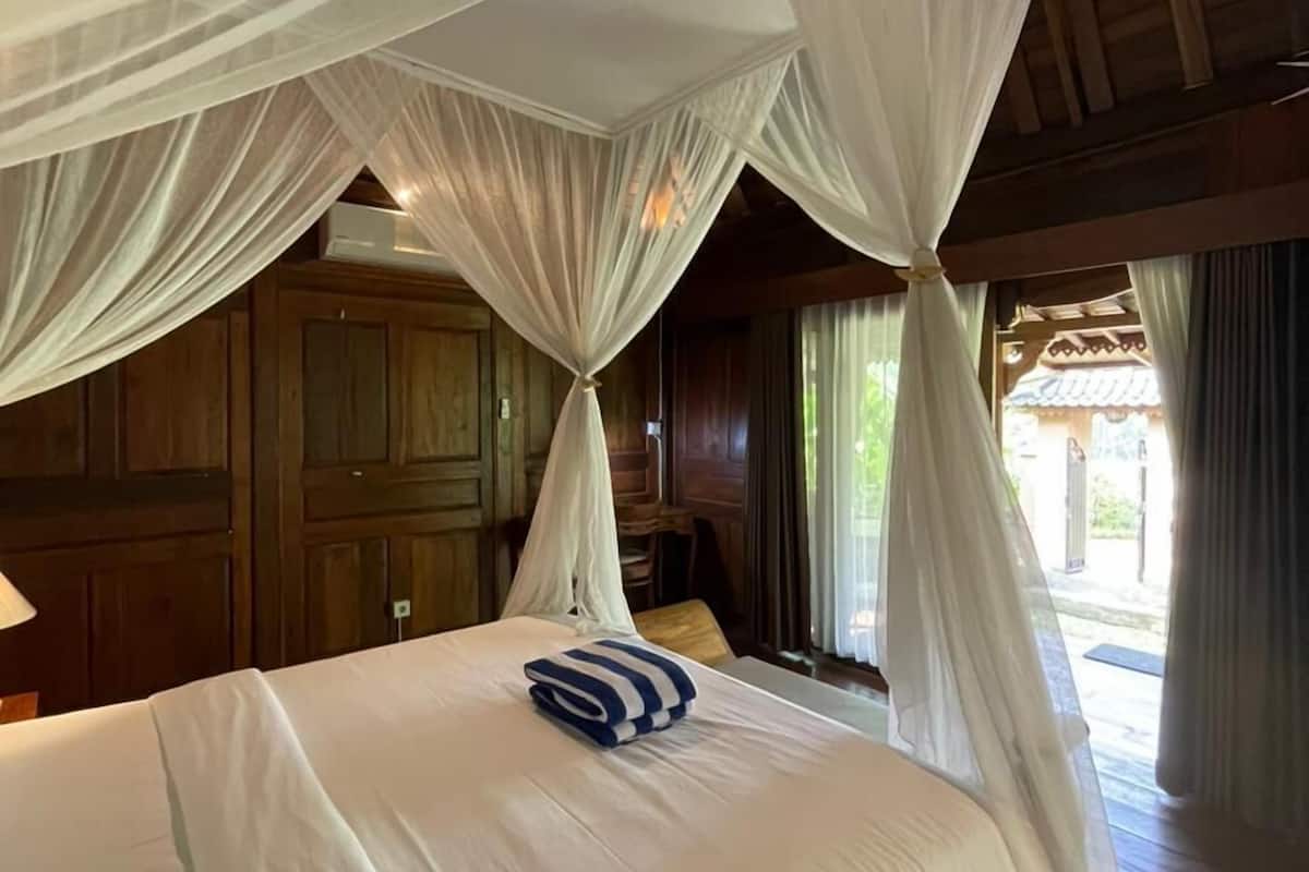 Una stanza con un letto ricoperto da lenzuola bianche e un baldacchino. Si nota un soffitto in legno e una porta sullo sfondo.
