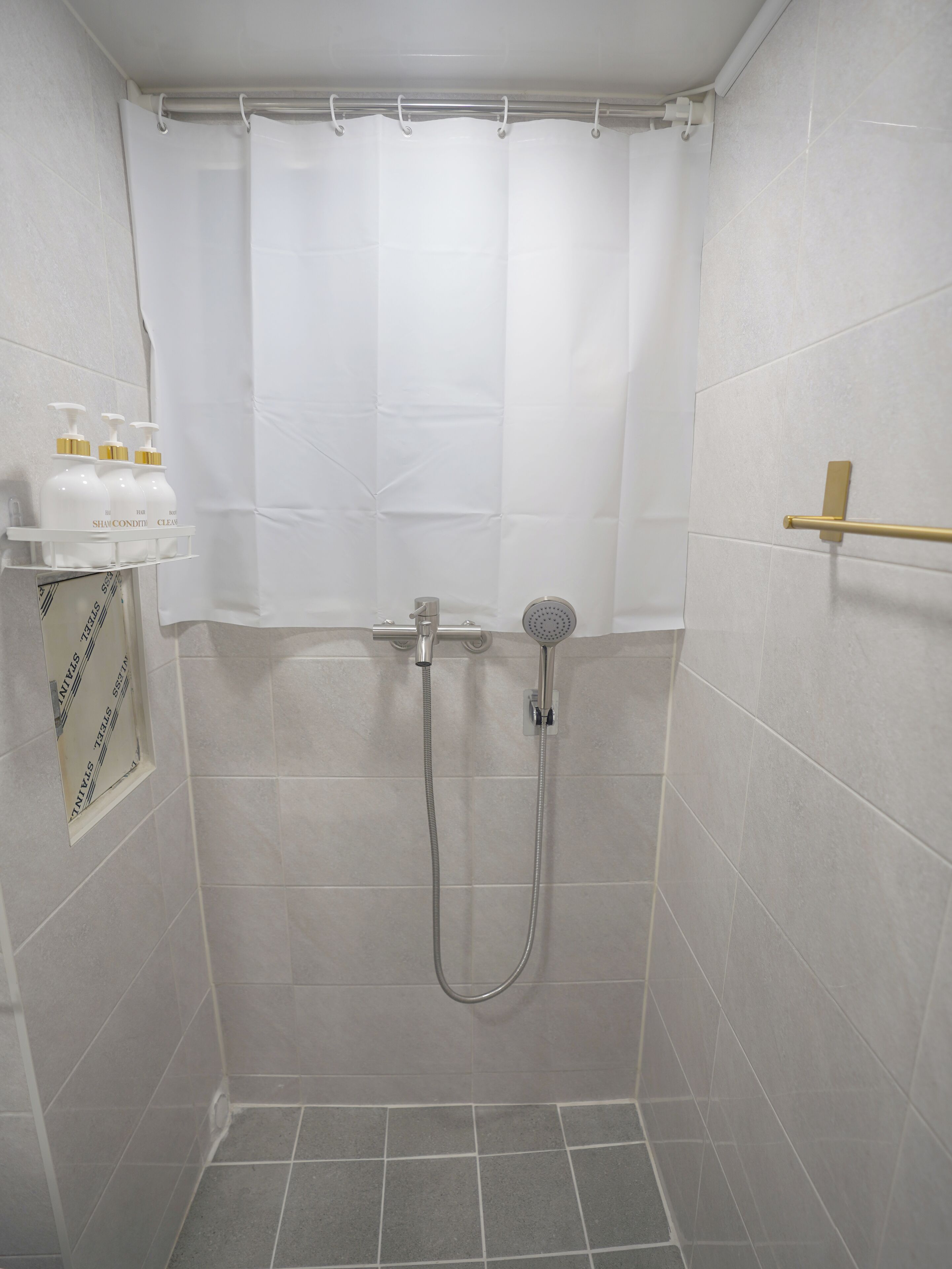 Eine Dusche mit Duschbrause, Wandarmatur und Duschvorhang. Auf einem Regal links befinden sich drei Flaschen Shampoo und Conditioner.