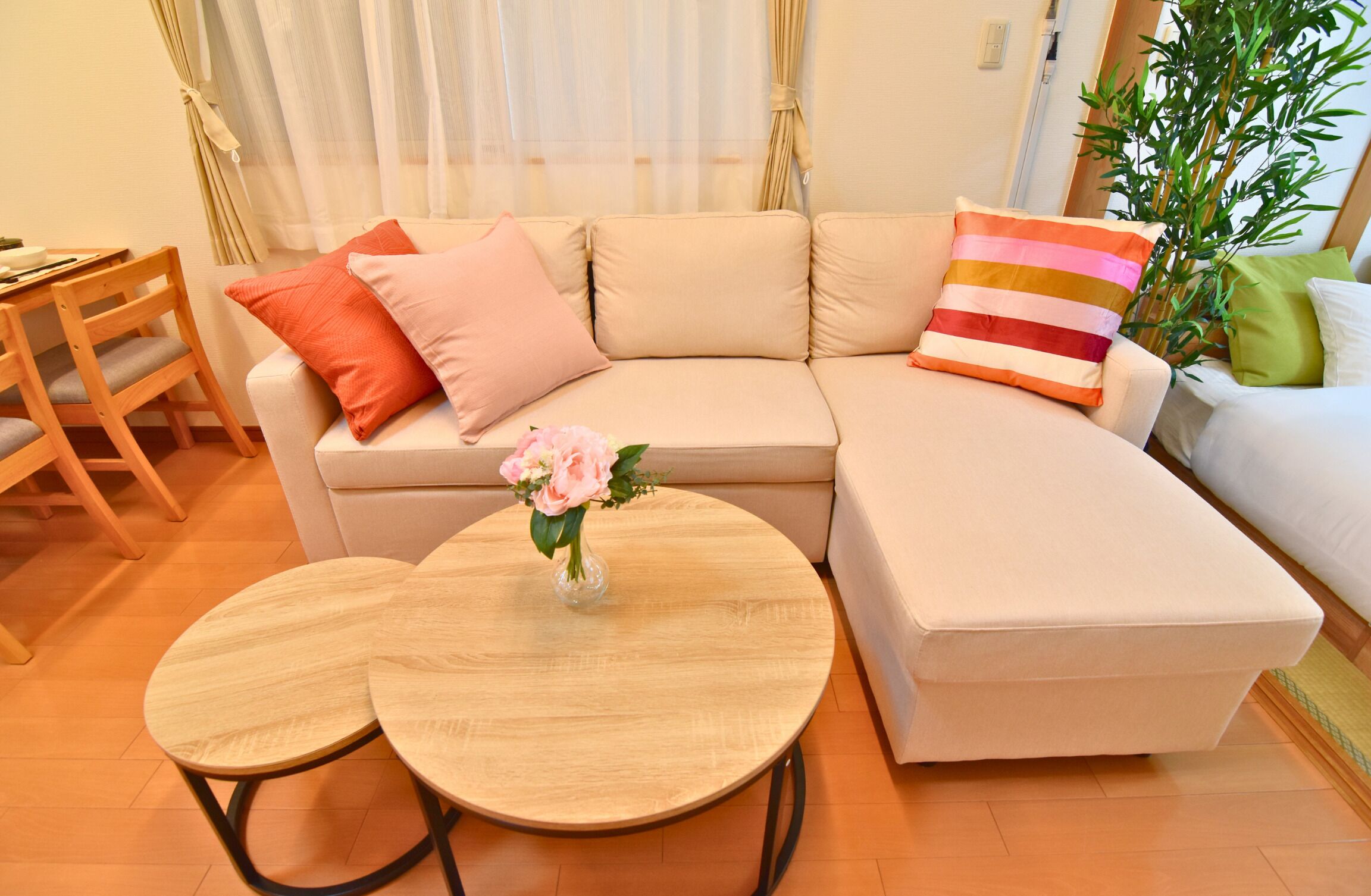 A living room with a beige sofa, two round wooden tables, a small wooden chair, and a vase with flowers.