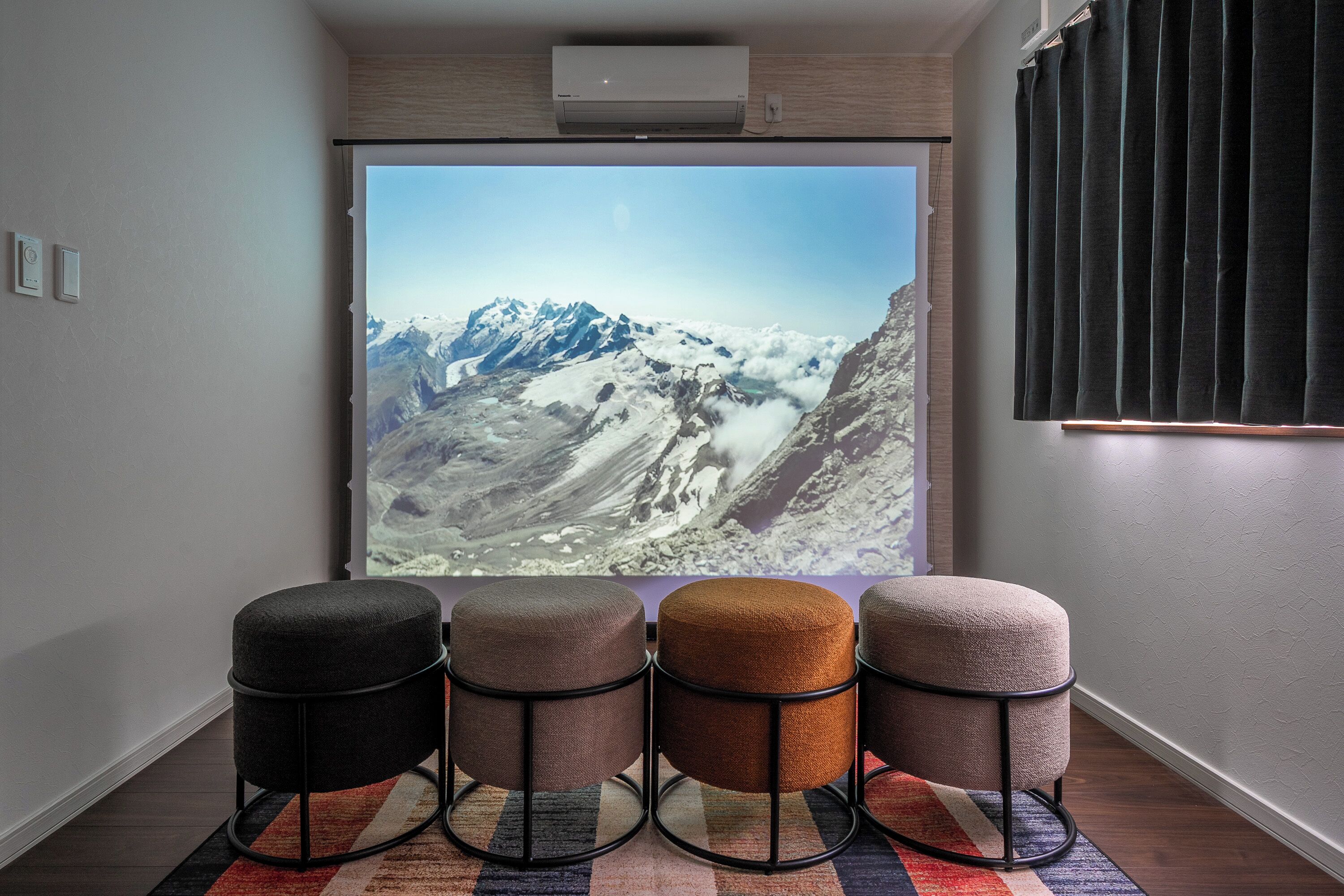 A room with a large window displaying a mountainous landscape, four ottomans in different colors, and a patterned rug.