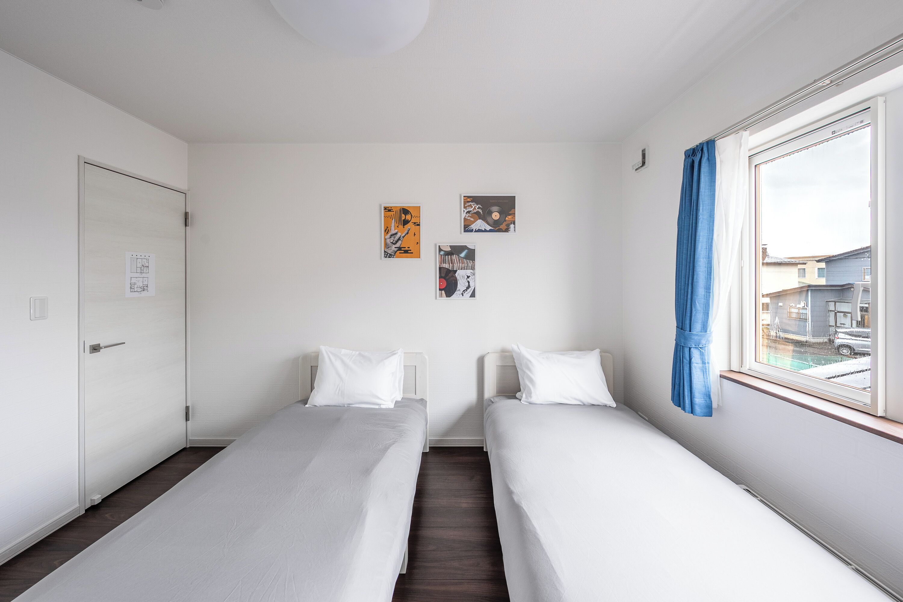 A room with two single beds, a door, and a window with a view of buildings.