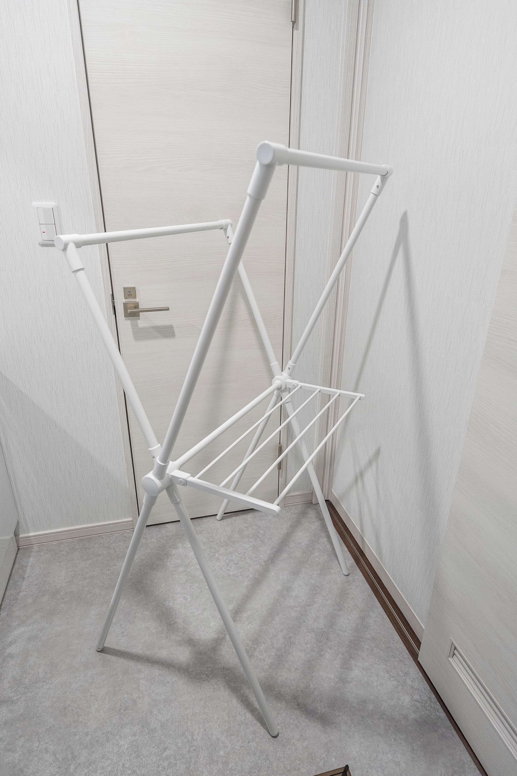 Un tendedero metálico blanco en un rincón de una habitación con alfombra clara y una puerta blanca.