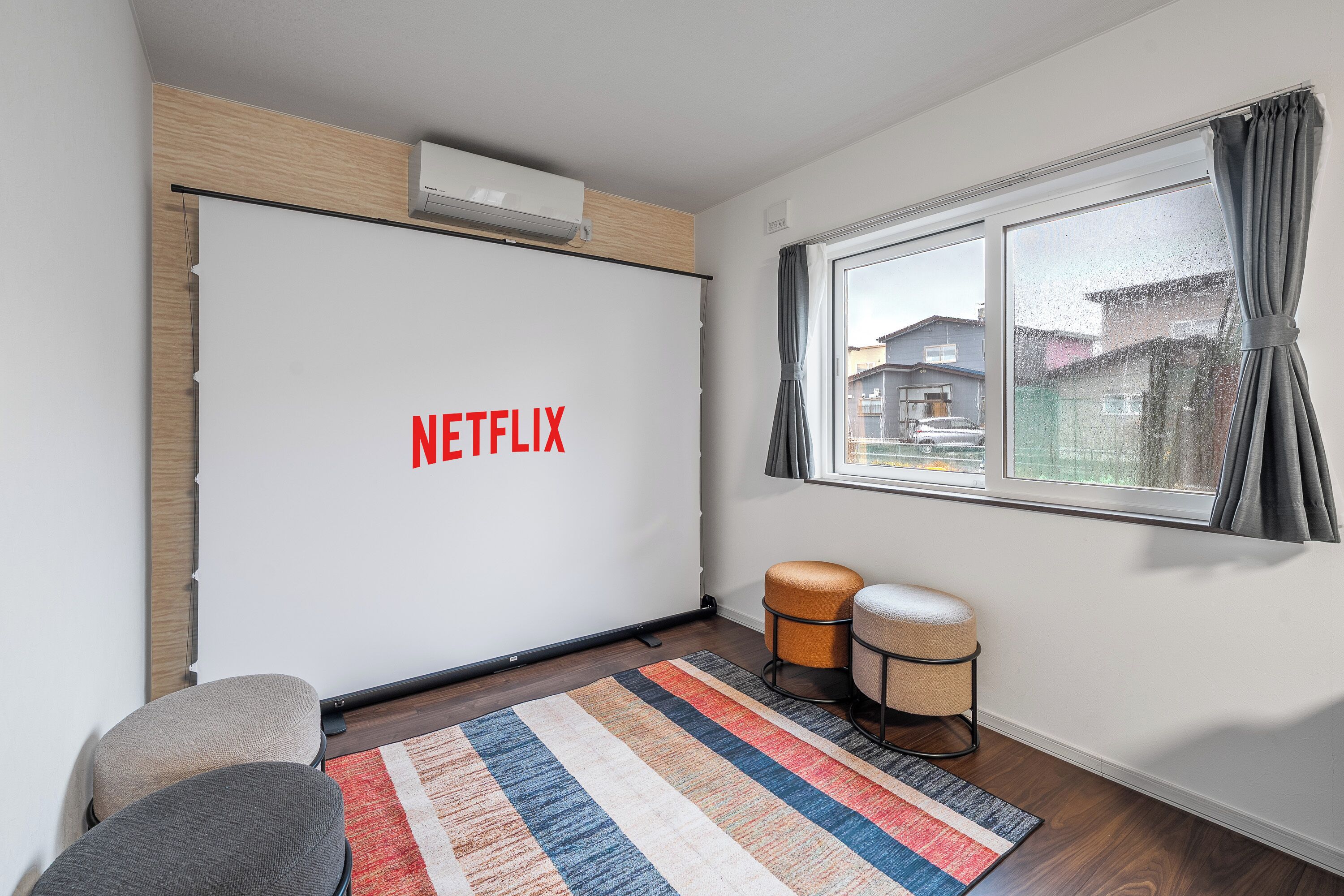 Um quarto com uma parede branca grande exibindo o logotipo da Netflix, uma janela com cortinas, um banquinho pequeno e um tapete listrado.