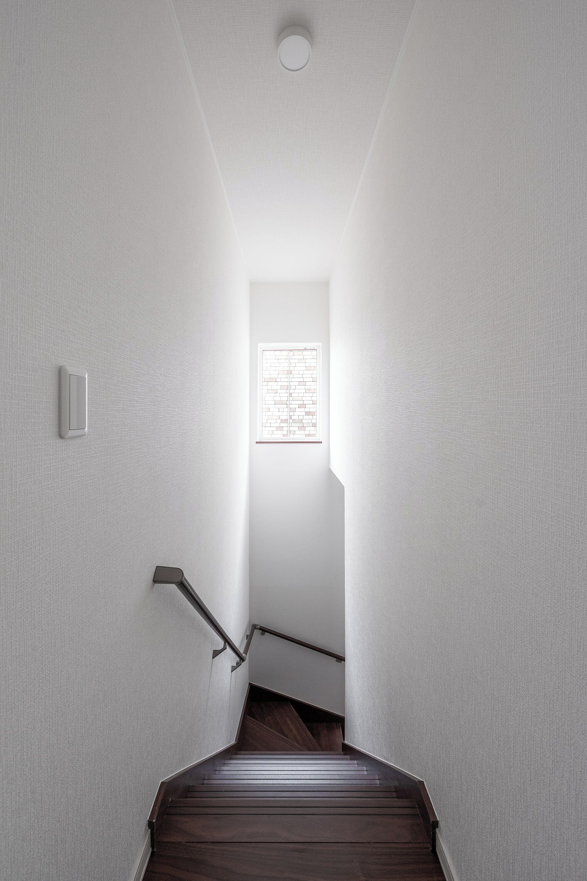 A narrow, well-lit hallway with a wooden staircase leading to a room with a window.