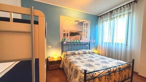 Een slaapkamer met een stapelbed, een eenpersoonsbed, een nachtkastje, een lamp, een raam met gordijnen en een spiegel aan de muur.