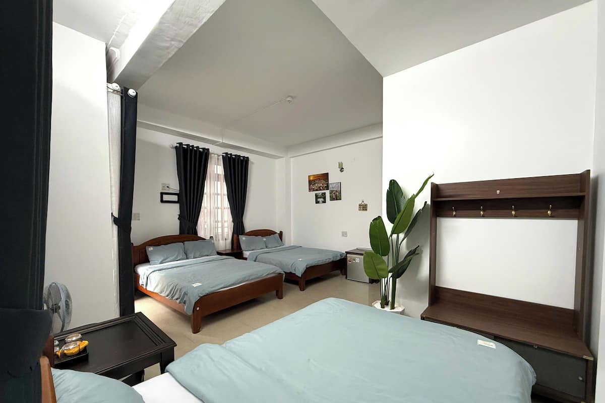 A hotel room with two single beds, a large bed, a bedside table with a lamp, a plant, and a wall-mounted TV.