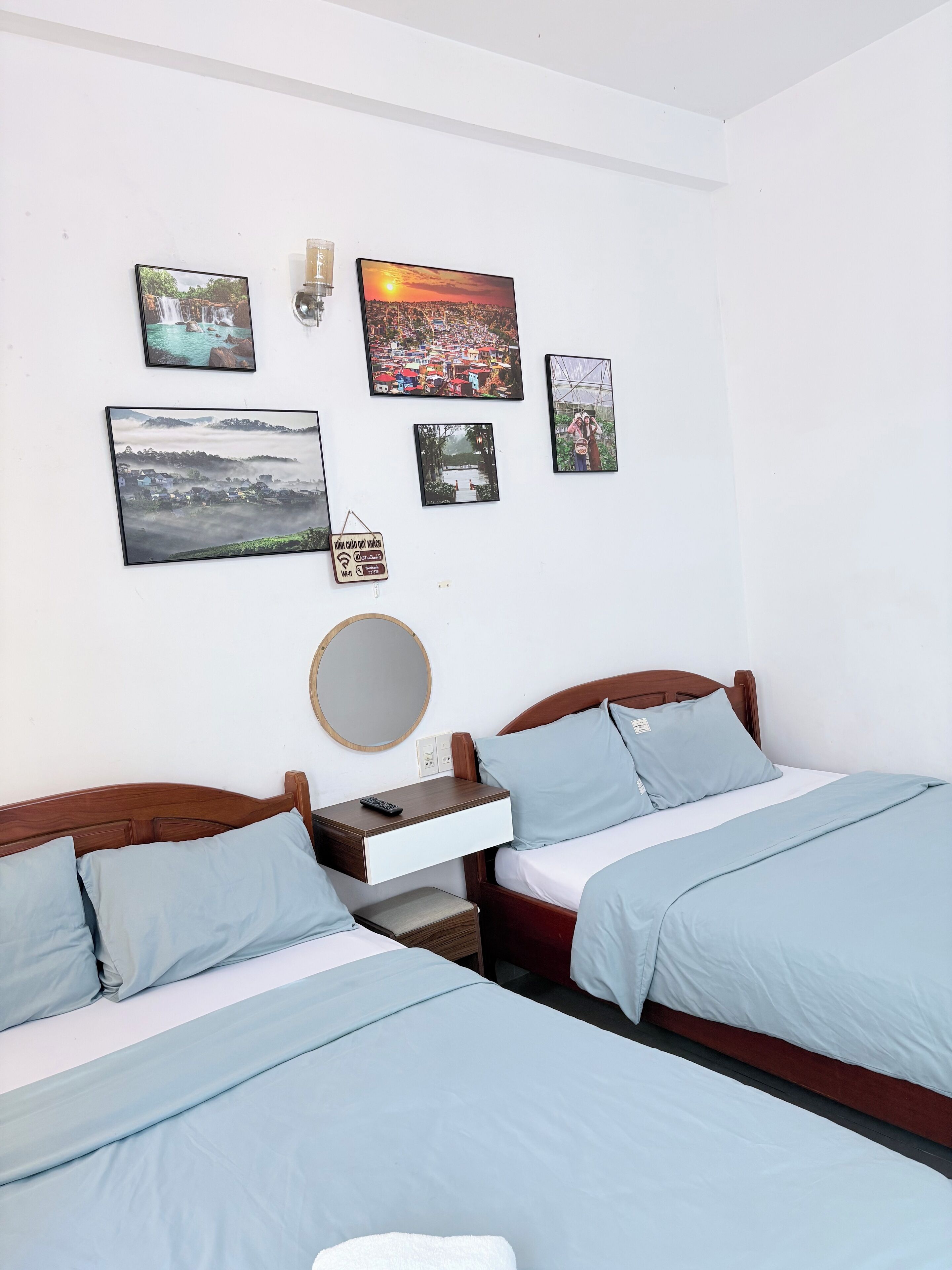 Habitación de hotel con dos camas, una mesita de noche con control remoto, un espejo montado en la pared y varias fotografías enmarcadas.