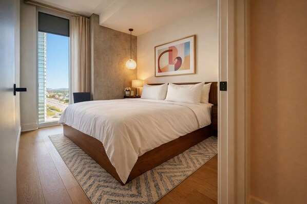 A hotel room with a large bed, a nightstand with a lamp, a view through a window, and a wall-mounted abstract art piece. - Skyline Views Rooftop Infinity Pool Rainey St (Austin)