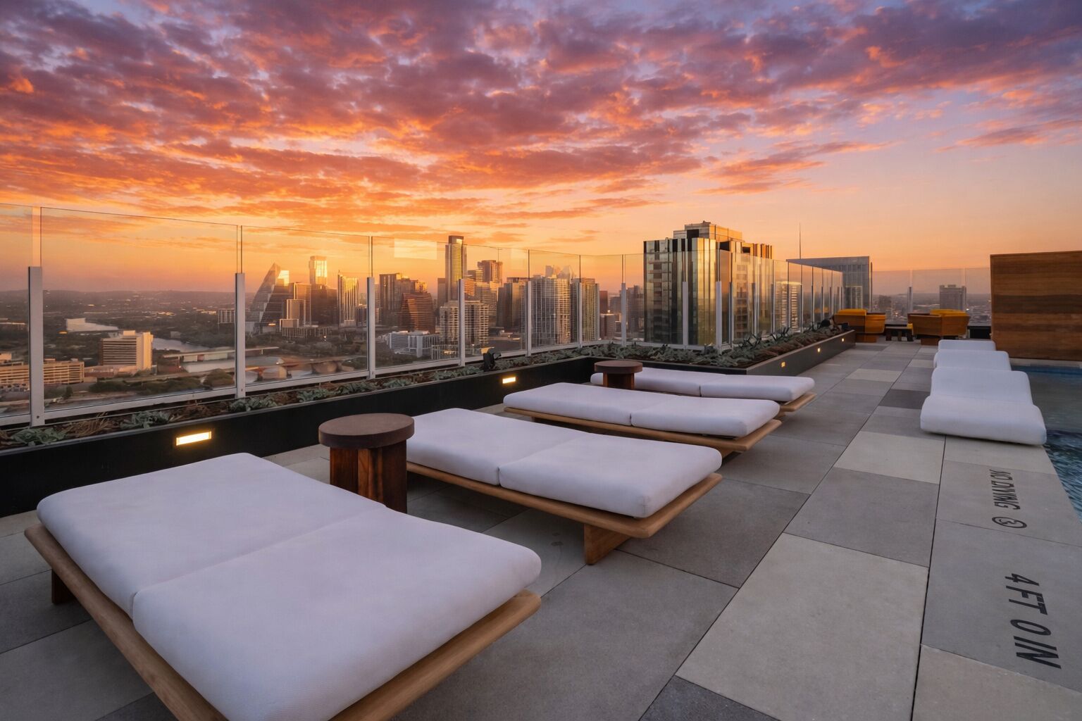 A rooftop lounge with white cushioned seating, a pool, and a city skyline view at sunset.