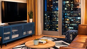 Ein modernes Wohnzimmer mit großem Fernseher, einem blauen Schrank, einer Pflanze und Blick auf die Stadt durch das Fenster.